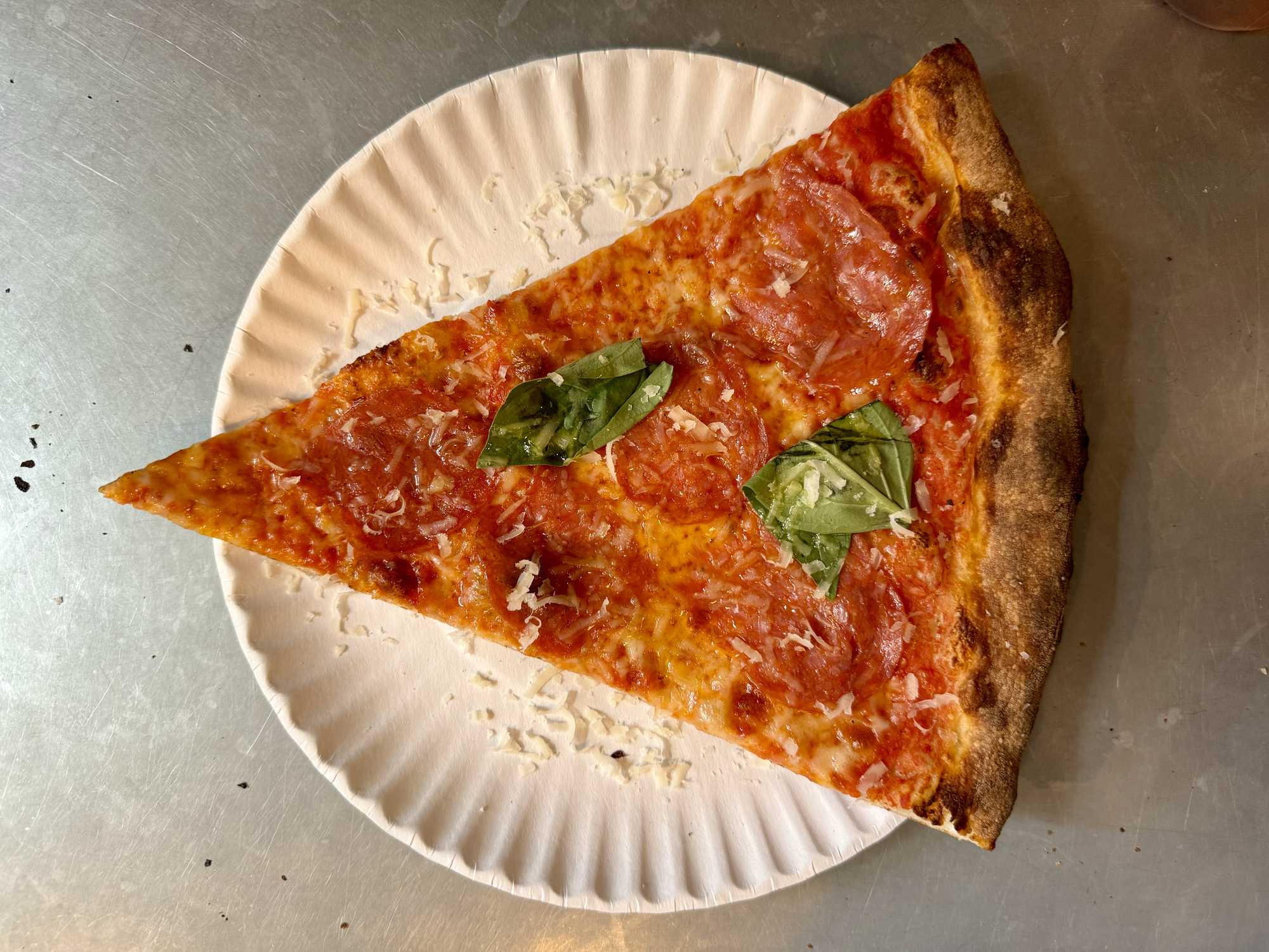 Top-down close-up of a triangular slice of thin-crust pizza with spicy sopressata and basil leaf on the metal counter at a pizzeria in the West Village, New York City