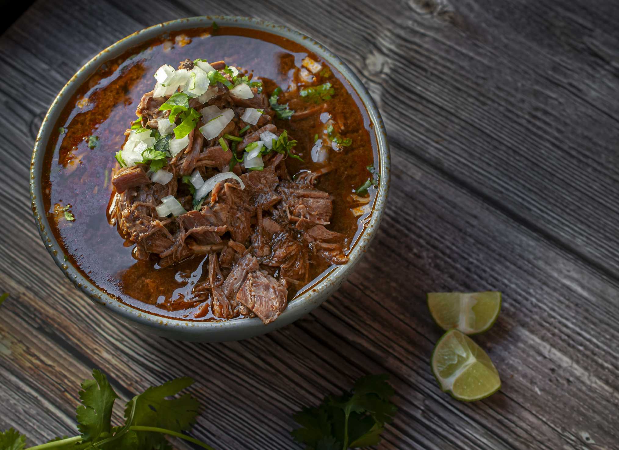 A bowl of BIRRIA or BARBACOA, a traditional Mexican dish with lemons and cilantro on the side over a wooden table, horizontal shot