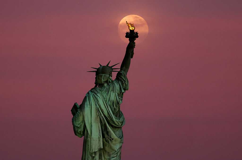 JERSEY CITY, NJ - MAY 11: The Flower Moon rises through clouds behind the Statue of Liberty as the sun sets in New York City on May 11, 2025, as seen from Jersey City, New Jersey.  (Photo by Gary Hershorn/Getty Images)